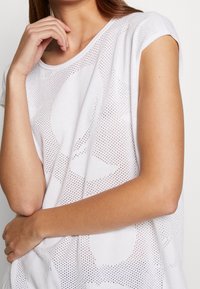 Bílé pletené tričko s volným střihem, vyznačující se texturovaným vzorem a krátkými rukávy, zdůrazňující jeho prodyšný a lehký materiál.