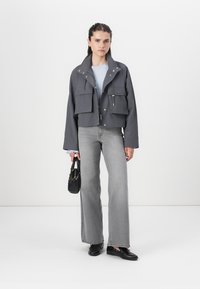 Kappet grå jakke med to frontlommer, knapp- og glidelås-lukking, kombinert med lys blå genser, grå vide jeans og svarte sko.