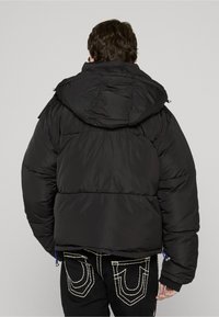 Črna puhasta jakna s kapuco, z rebrastim vzorcem in elastičnimi manšetami. Izstopajoči detajli vključujejo vrvico na spodnjem robu in stranski žep.