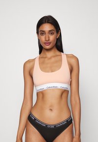 Barack színű sportmelltartó széles fehér pánttal, amelyen a Calvin Klein logo található; fekete bikini stílusú alsóval párosítva, amelyen szintén található a logópánt.