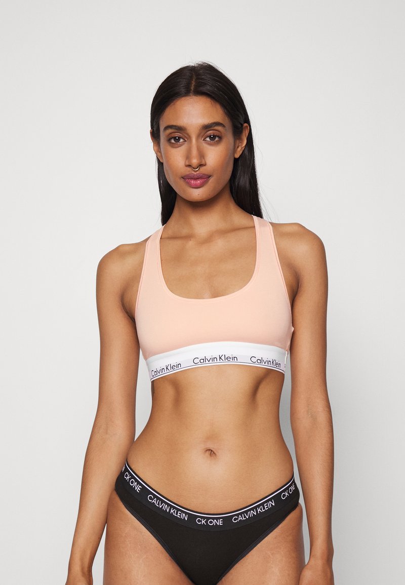 Barack színű sportmelltartó széles fehér pánttal, amelyen a Calvin Klein logo található; fekete bikini stílusú alsóval párosítva, amelyen szintén található a logópánt.