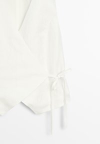 Zbliżenie na biały przewiązywany strój z bocznym wiązaniem, wykonany z lekkiego materiału na jednolitym tle.