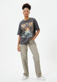 T-shirt oversize grigio con un design grafico celestiale colorato, abbinato a pantaloni cargo beige chiaro e sneakers bianche.