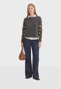 Grijze wollen trui met gestreepte groene en beige mouwen, gecombineerd met donkere denim wijde broek. Geaccentueerd met een bruine handtas.