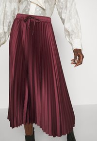 Gonna midi plissettata bordeaux con vita elastica e coulisse, realizzata in tessuto liscio e lucido, caratterizzata da pieghe verticali nette.