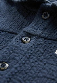 Navyfarbendes, strukturiertes Hemd mit Muster, ausgestattet mit einem Knopf, der "Ted Baker" beschriftet ist, und einem flachen Kragen. Eine Nahaufnahme zeigt die Stoffdetails.