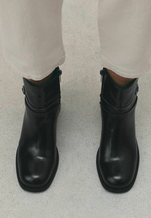Black leather ankle boots with square toes worn with light-colored pants on a light gray floor surface.