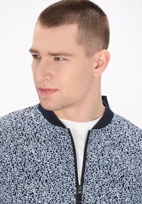 Giacca bomber navy con motivo astratto bianco e colletto nero; presenta una zip frontale e una texture di tessuto morbido. T-shirt bianca sottostante.