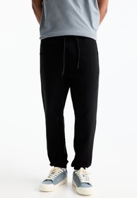 Črne joggers hlače z vrvico v pasu, oblikovane z ozkimi nogami in stranskimi žepi, v kombinaciji z svetlo modrimi športnimi copati.