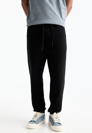 Schwarze Jogginghose mit verstellbarem Bund, schmal zulaufendem Beinschnitt und seitlichen Taschen, kombiniert mit hellblauen Turnschuhen.