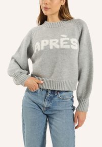 GrauerStrickpullover mit geripptem Saum und Bündchen, auf der Brust mit dem Wort "APRÈS" in Weiß versehen. Kombiniert mit blauen Jeans.