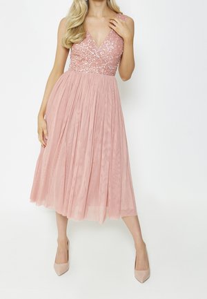 Femme portant une robe sans manches rose à paillettes avec une jupe en tulle plissée et des talons hauts couleur nude, posant devant un fond blanc.
