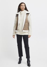 Brun mockaskinnsväst med krämfärgade faux fur-krage, kombinerad med en krämfärgad kabelstickad tröja, svarta jeans och svarta ankelboots.