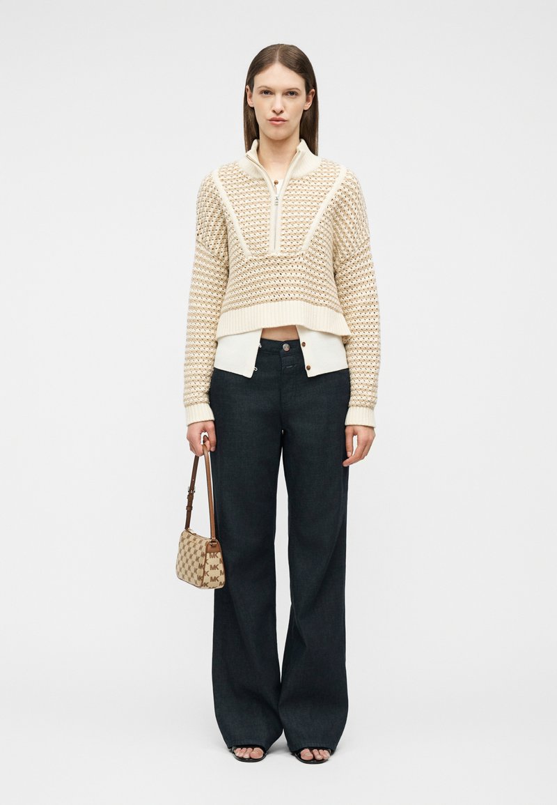 Γυναίκα που φοράει μπεζ υφασμάτινο πουλόβερ με φερμουάρ, κρεμ πουκάμισο με κουμπιά από κάτω, μαύρο παντελόνι φαρδιάς γραμμής, μαύρα σανδάλια και κρατάει μια μικρή μπεζ τσάντα με σχέδια.