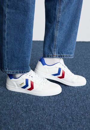 Baskets blanches avec des bandes chevron bleu et rouge portées avec des chaussettes blanches et un jean bleu, debout sur un sol moquetté bleu.