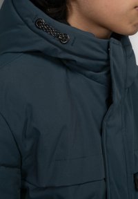 Mörkt turkos vadderad jacka med hög krage, med en texturerad huva och snoddar, visad från sidan.