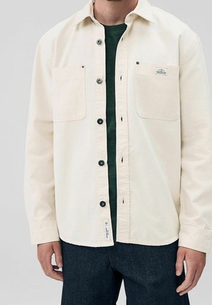 Mężczyzna w kremowej koszuli na guziki nałożonej na ciemnozielony t-shirt z dwoma kieszeniami na piersi i ciemnoniebieskich dżinsach, ręce swobodnie opuszczone wzdłuż ciała.