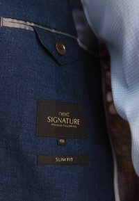 Tamsiai mėlynos spalvos kostiumo švarkas, kurio viduje matyti etiketė su užrašu "next SIGNATURE PREMIUM TAILORING" bei informacija apie dydį (40R) ir pritaikymą (SLIM FIT).