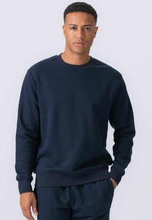 Mężczyzna stojący z jedną ręką w kieszeni, ubrany w gładką, ciemnogranatową bluzę i dopasowane spodnie na białym tle.