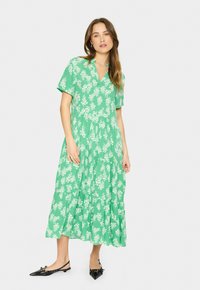 Rochie midi verde cu flori, cu mâneci scurte, volane în straturi și decolteu în V. Purtată cu pantofi plata negri cu vârf ascuțit, accentuați cu fundițe.