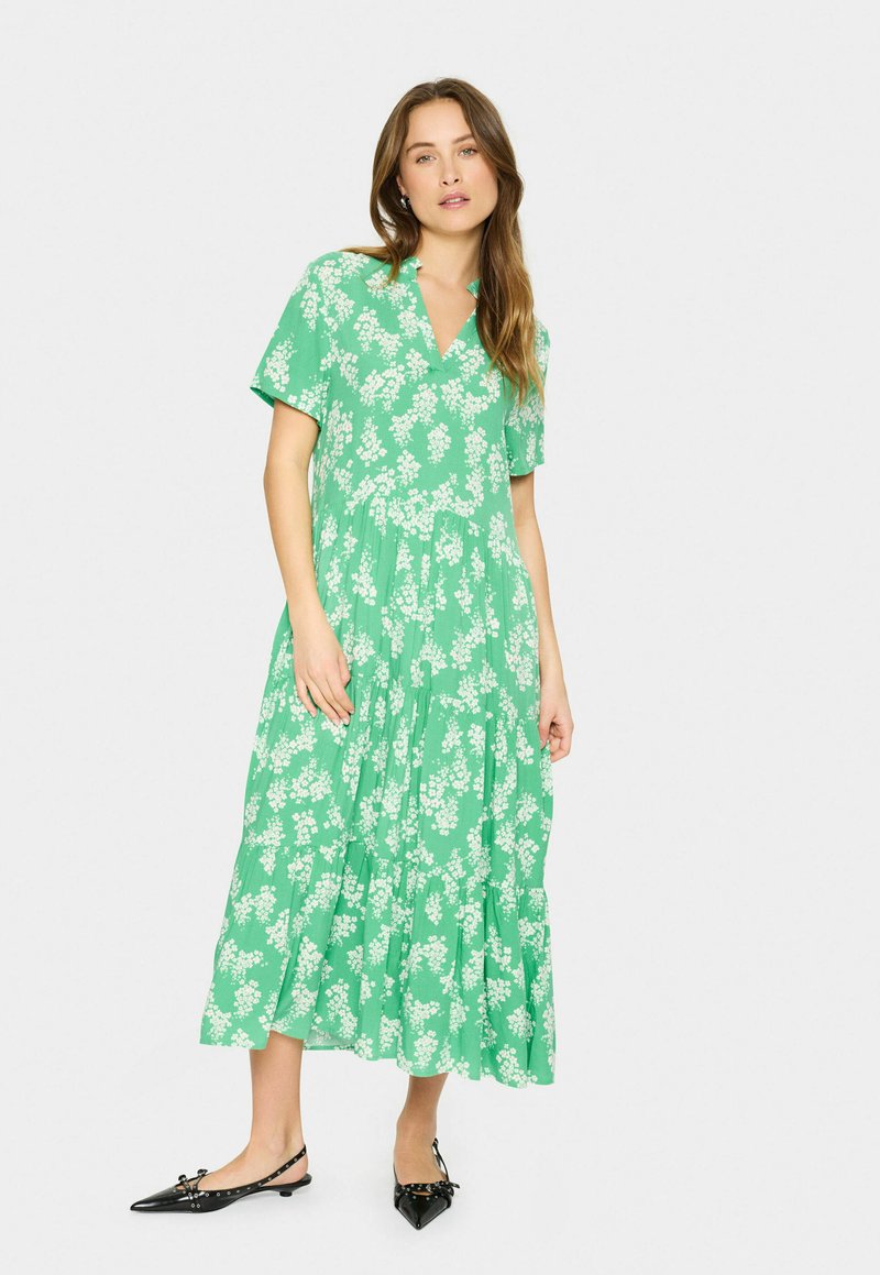 Rochie midi verde cu flori, cu mâneci scurte, volane în straturi și decolteu în V. Purtată cu pantofi plata negri cu vârf ascuțit, accentuați cu fundițe.
