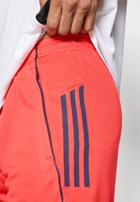 Červené športové šortky s tmavomodrými bočnými pruhmi a priedušnou tkaninou. Pás je zrejme elastický a na okrajoch je viditeľné prešívanie.