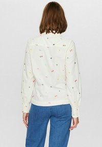 Camicia color crema con motivi floreali e animali ricamati, colletto con bottoni, maniche lunghe con polsini elastici, abbinata a pantaloni blu.