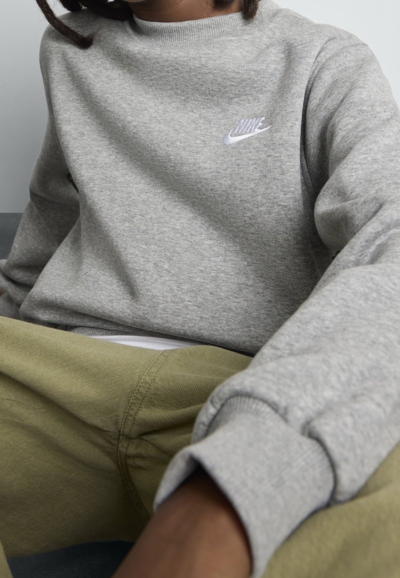 Personne portant un sweat-shirt gris Nike et un pantalon vert olive, assise avec les bras reposant vers l'avant.