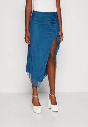 Femme portant une jupe midi bleue froncée avec une fente latérale haute et des talons compensés blancs, debout contre un mur clair uni.