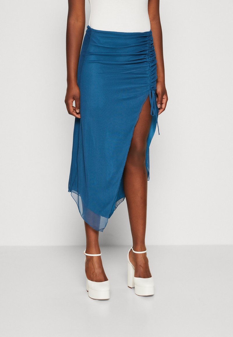 Femme portant une jupe midi bleue froncée avec une fente latérale haute et des talons compensés blancs, debout contre un mur clair uni.