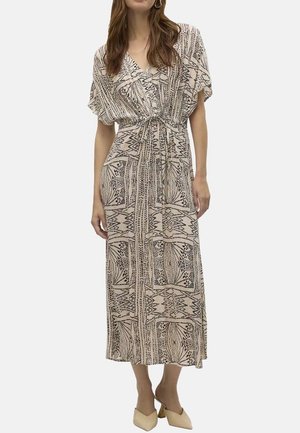 Femme portant une robe portefeuille à manches courtes beige et noire à motifs, nouée à la taille, debout dans des chaussures pointues de couleur claire.