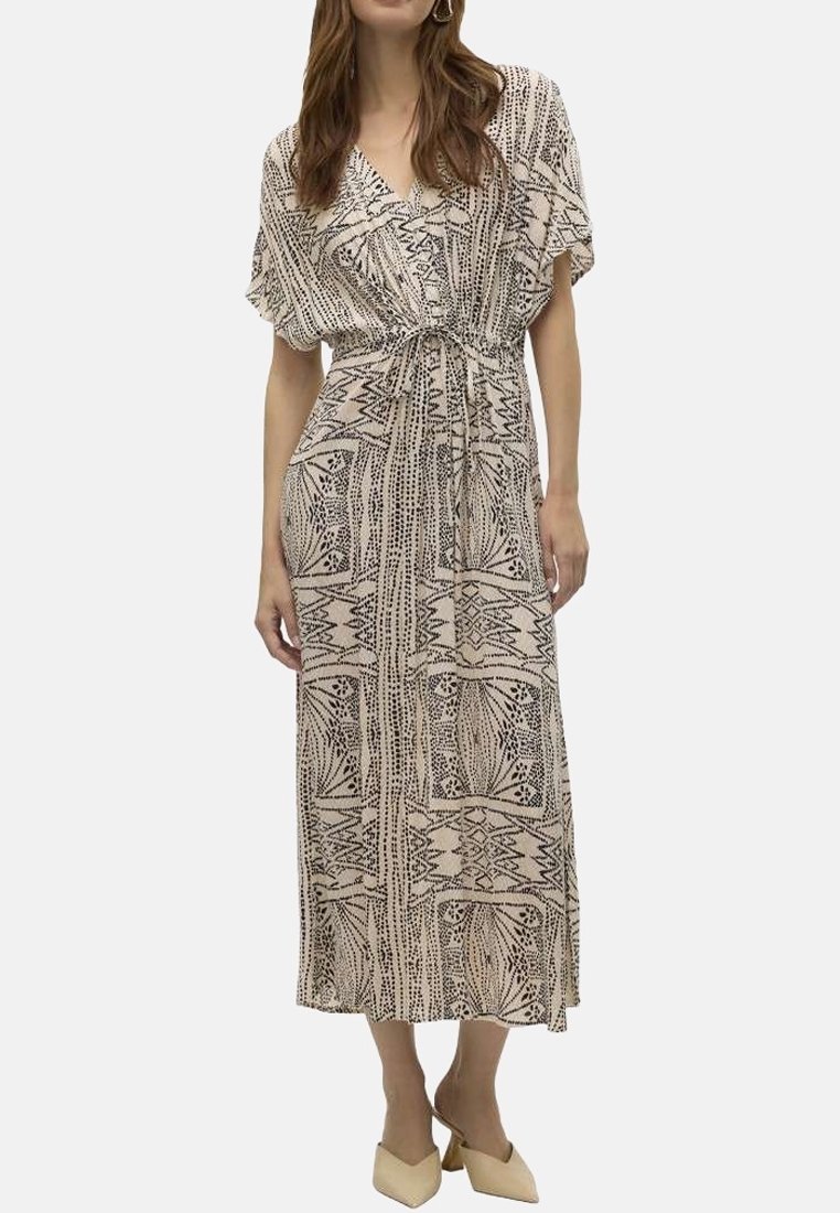 Femme portant une robe portefeuille à manches courtes beige et noire à motifs, nouée à la taille, debout dans des chaussures pointues de couleur claire.
