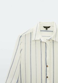 Blouse rayée avec des rayures verticales blanches et bleues, en tissu léger. Dotée d'un col pointu et de boutons marron sur le devant.
