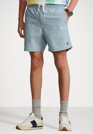 Mann trägt hellblaue Shorts mit Kordelzug, beige-schwarze Turnschuhe, graue Socken und eine Armbanduhr, steht mit den Händen in den Taschen.