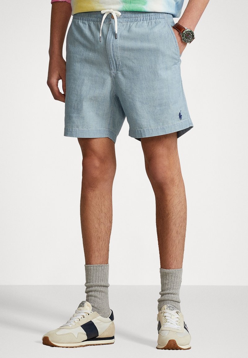 Homme portant un short bleu clair à cordon, des baskets beige et noires, des chaussettes grises et une montre au poignet, debout les mains dans les poches.