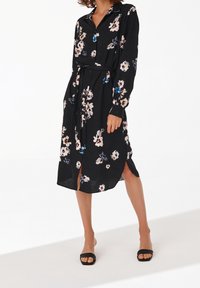 Černé květinové šaty vyrobené z lehké tkaniny, s knoflíkovým zapínáním vpředu, dlouhými rukávy a pásem v pase. Jsou zde boční rozparky.