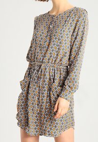 Sukienka z długim rękawem z wzorzystej tkaniny, prezentująca geometryczny wzór w diamenty w kolorach niebieskim, pomarańczowym i żółtym. Posiada ściągacz w talii oraz guziki.