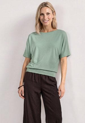 Frau mit schulterlangem blondem Haar, lächelnd, trägt ein lockeres hellgrünes Kurzarmtop und dunkelbraune Hosen mit weitem Bein.
