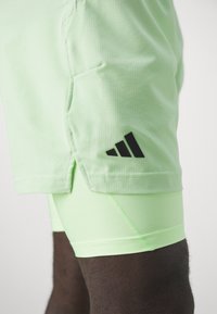 Shorts de sport vert clair avec une surface texturée, arborant un logo Adidas noir sur le côté, associés à un short de compression vert assorti en dessous.