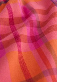 Prim-plan al unui material cu o textură moale, având un model în carouri portocalii, roz și roșii, cu dungi suprapuse și pliuri.