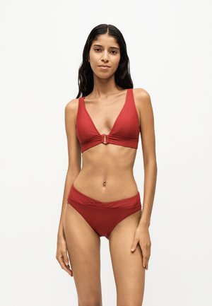 Donna con capelli lunghi e scuri che indossa un bikini rosso intenso con top a V e slip con fascia piegata, in piedi davanti a uno sfondo bianco semplice.