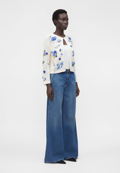 Kvinna som bär en vit stickad kofta med blå blomstermönster, vit topp och vida blå jeans, stående mot en enfärgad bakgrund.
