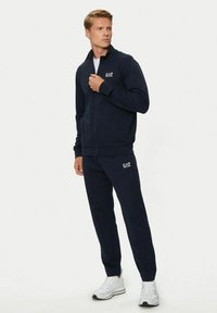 Completo da ginnastica blu navy realizzato in tessuto morbido, composto da una giacca con zip e pantaloni coordinati. Logo EA7 ricamato sulla giacca e sui pantaloni.