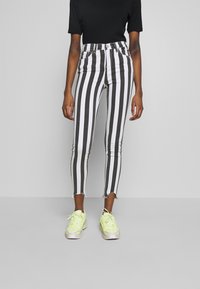 Mujer vistiendo jeans skinny de rayas verticales en blanco y negro de cintura alta con dobladillos deshilachados y zapatillas de neón amarillo contra un fondo gris.