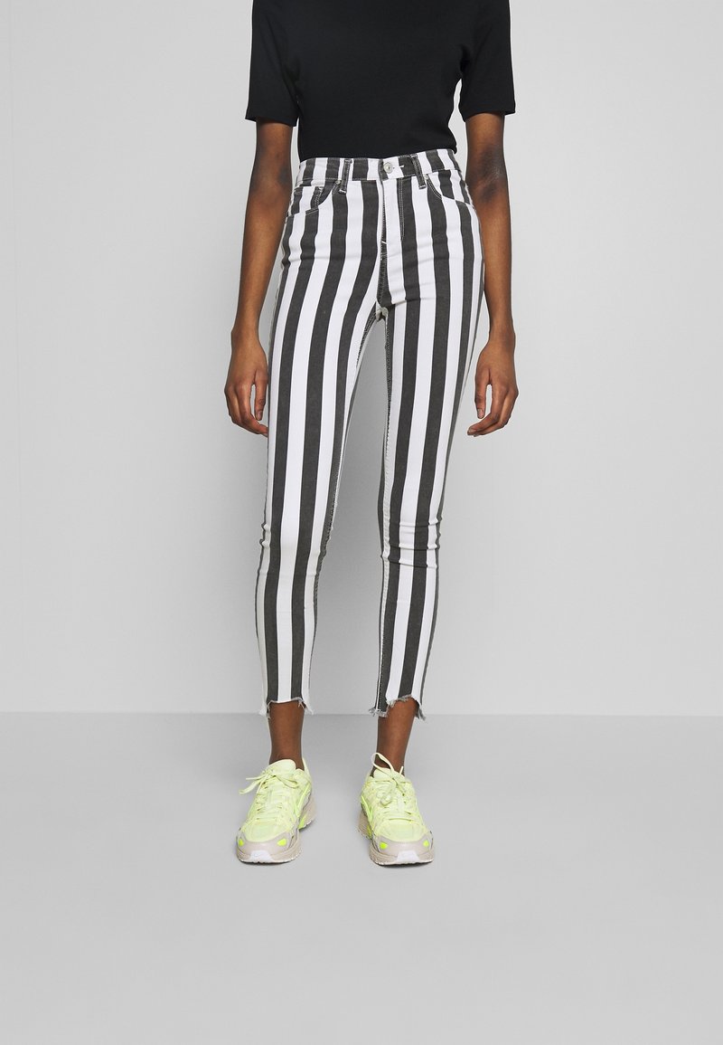 Mujer vistiendo jeans skinny de rayas verticales en blanco y negro de cintura alta con dobladillos deshilachados y zapatillas de neón amarillo contra un fondo gris.