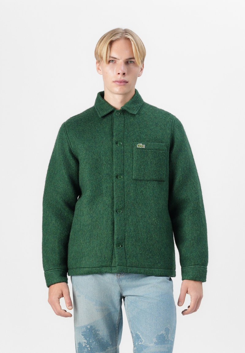 Veste en laine mélangée verte à boutons avec une poche poitrine, un col arrondi et une texture subtile. Présente un petit logo brodé sur la poche.