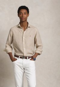 Hellbeige Leinenhemd mit Button-Down-Kragen, hochgekrempelten Ärmeln, kombiniert mit weißen Hosen und einem braunen Leder Gürtel. Die lässige Armbanduhr ist sichtbar.