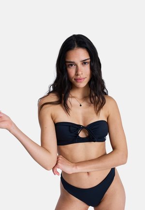 Kobieta z długimi ciemnymi włosami, ubrana w czarne, bezramionowe bikini z kokardą z przodu i naszyjnikiem z zawieszką, stojąca na jednolitym jasnym tle.