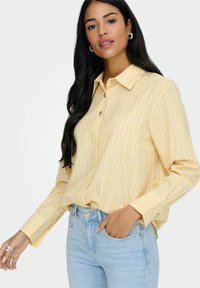 Chemise jaune à boutons avec des rayures verticales bleues, manches longues et col ; portée avec un jean ajusté bleu clair.