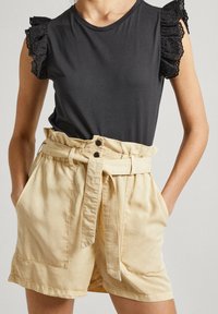 Czarna koszula z krótkim rękawem ozdobionym falbankami i dziurkowanym wzorem, zestawiona z beżowymi, wysokim stanem szortami z paskiem do wiązania i bocznymi kieszeniami.